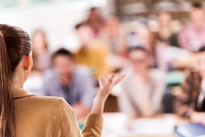 Rear view of female teacher giving a lecture in the classroom. There are people in the background.